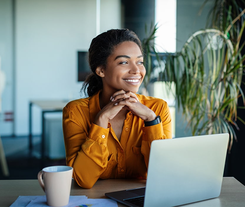 A woman in a yellow blouse sits at her laptop researching 2024 cybersecurity predicitons with a cup of coffee looking out the window. 