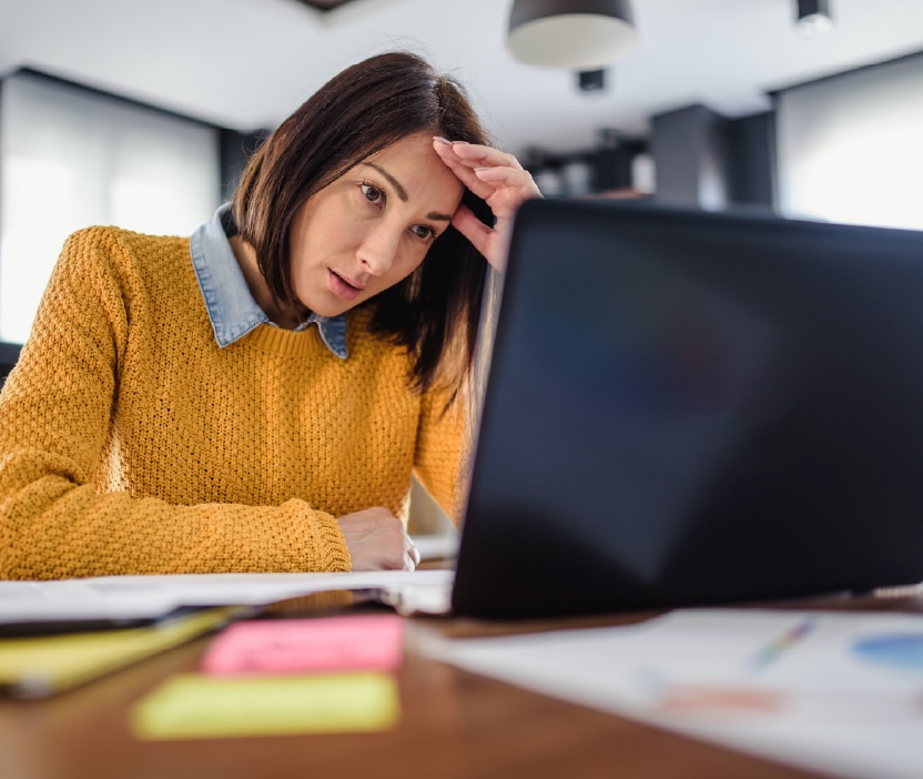 A woman wearing a yellow sweater looks defeated at a laptop with a blue screen of death.