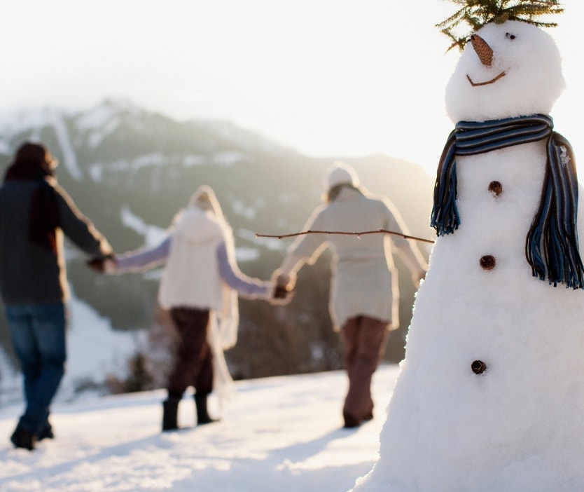 A couple hand in hand in the snow walking behind a dressed-up snowman.