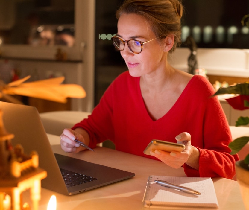 A woman in a red sweater sits at her laptop with her phone in one hand and her credit card in the other.