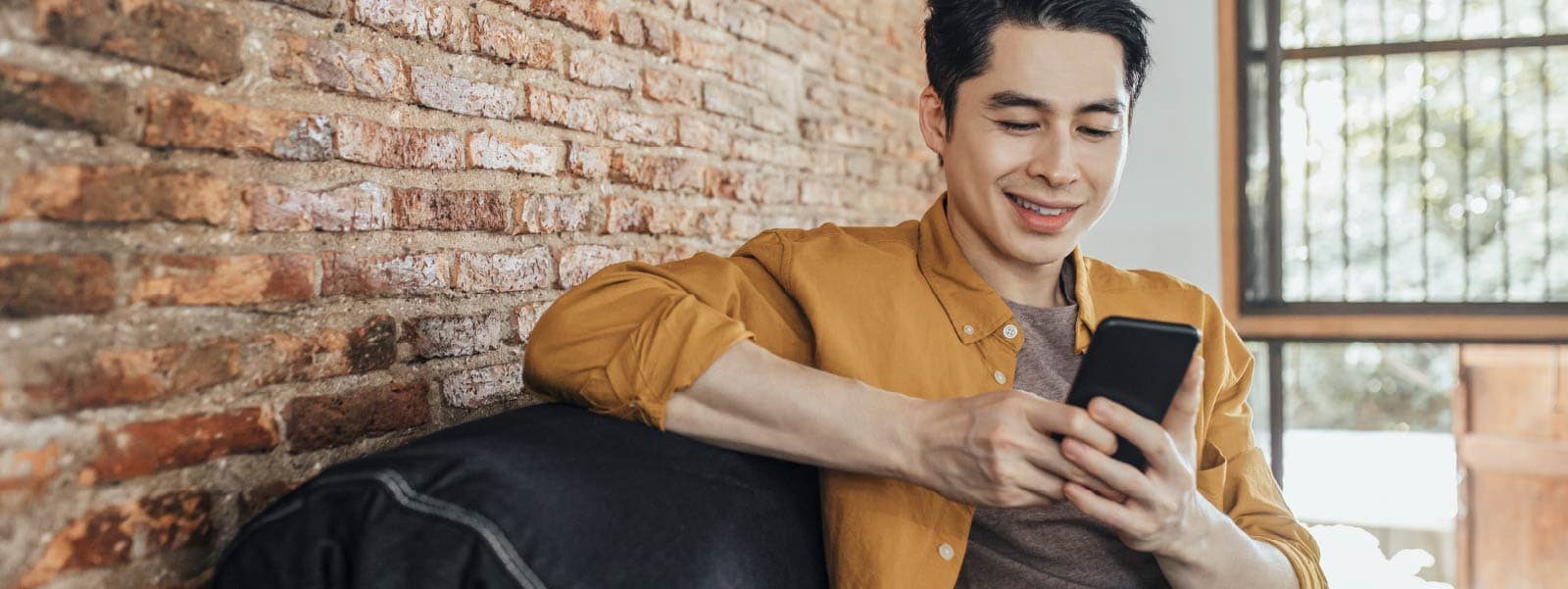 A guy who is unaware he was targeted in a doxxing attack sits in a mustard-yellow shirt on a bench in front of a brick wall, scrolling through his phone.