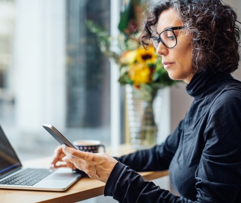 A woman with glasses sits at a table in front of a laptop while looking at her phone.