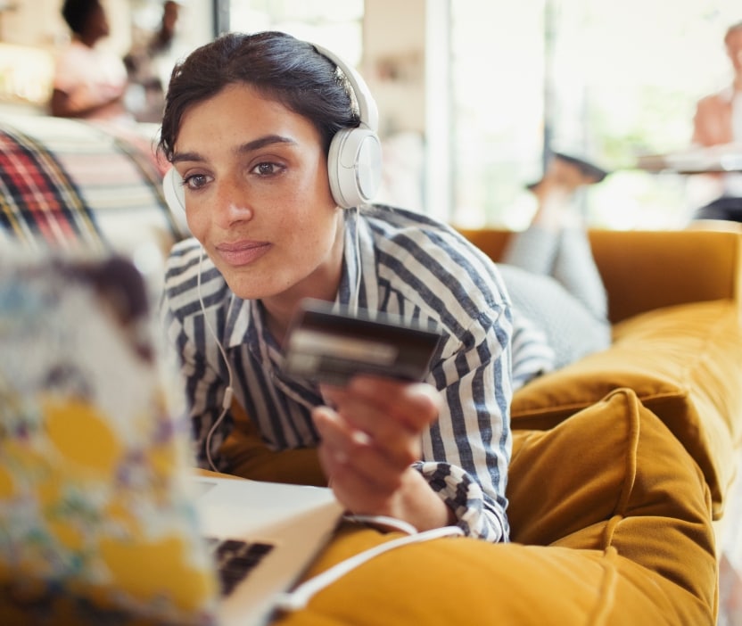 A woman lying down on a couch looks at her computer with a credit card in hand, falling for a gift card scam. 