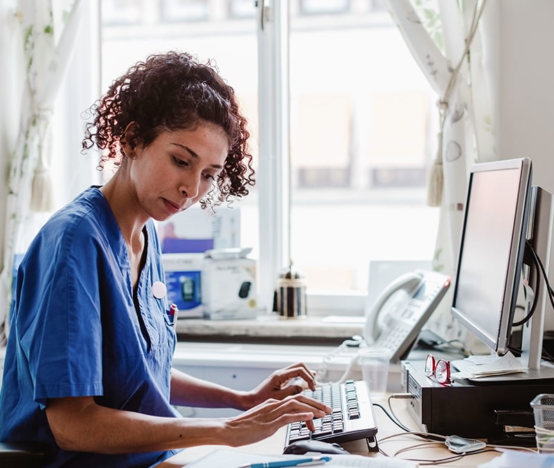 A healthcare professional wearing blue scrubs sits at a desk in a brightly lit room, typing on a keyboard while learning about hospital cyberattacks. A computer monitor, telephone, and medical supplies are visible on the desk.