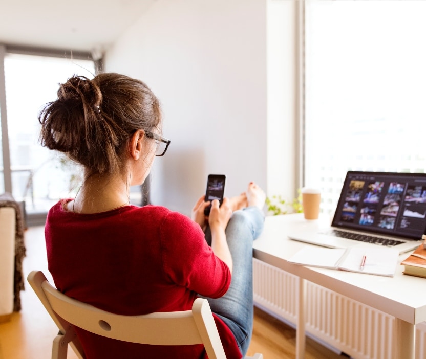 A woman sits with her feet up on a table with a laptop and a phone in front of her.