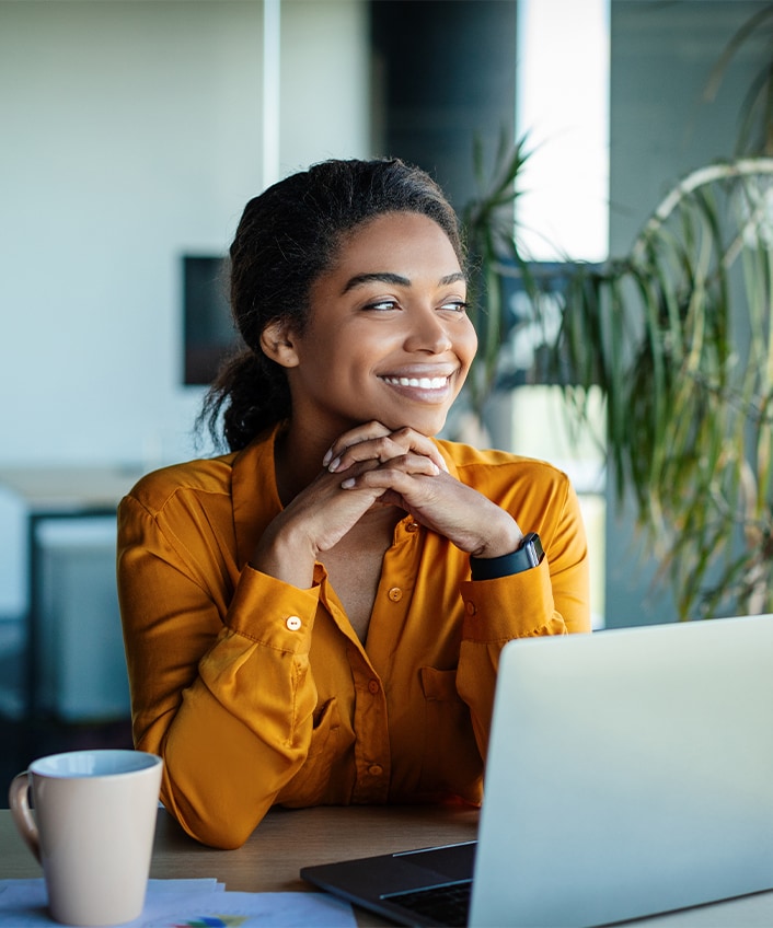 A woman in a yellow blouse sits at her laptop researching 2024 cybersecurity predicitons with a cup of coffee looking out the window. 