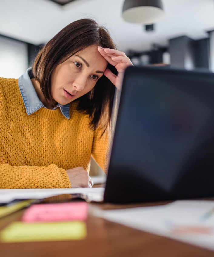 A woman wearing a yellow sweater looks defeated at a laptop with a blue screen of death.