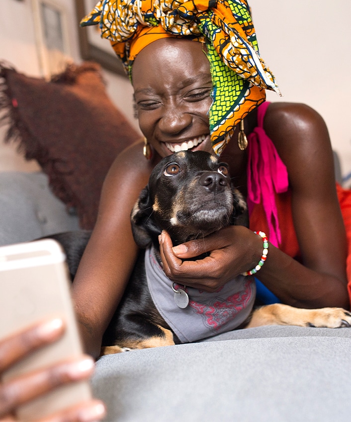 A woman checks looks on her phone while laying on a sofa with her dog, and she is learning about if pet tech can be hacked.