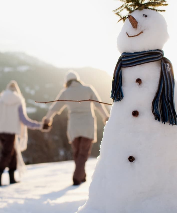 A couple hand in hand in the snow walking behind a dressed-up snowman.