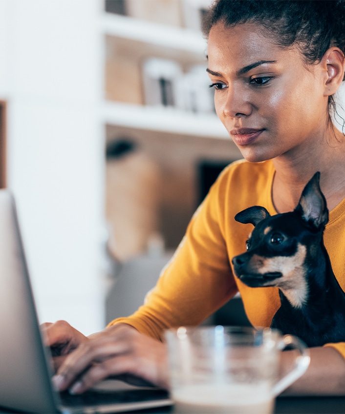 A woman sits at a table with her dog in her lap having brought her computer back from the dead. 