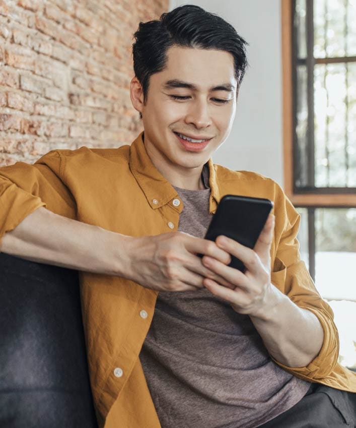 A guy who is unaware he was targeted in a doxxing attack sits in a mustard-yellow shirt on a bench in front of a brick wall, scrolling through his phone.