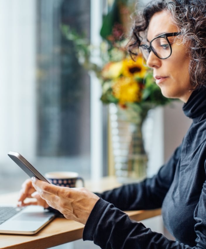 A woman with glasses sits at a table in front of a laptop while looking at her phone.