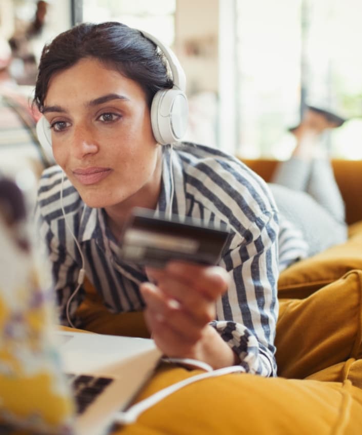 A woman lying down on a couch looks at her computer with a credit card in hand, falling for a gift card scam. 