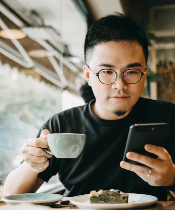 Man sitting in a coffee shop holding a mug of coffee and looking at his phone.