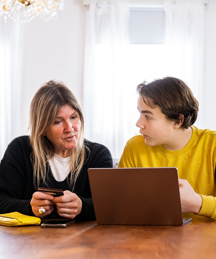 A woman sits at a table with her son looking at a laptop where they are encountering a scammer using loss aversion to influencer them to fall for a scam. 